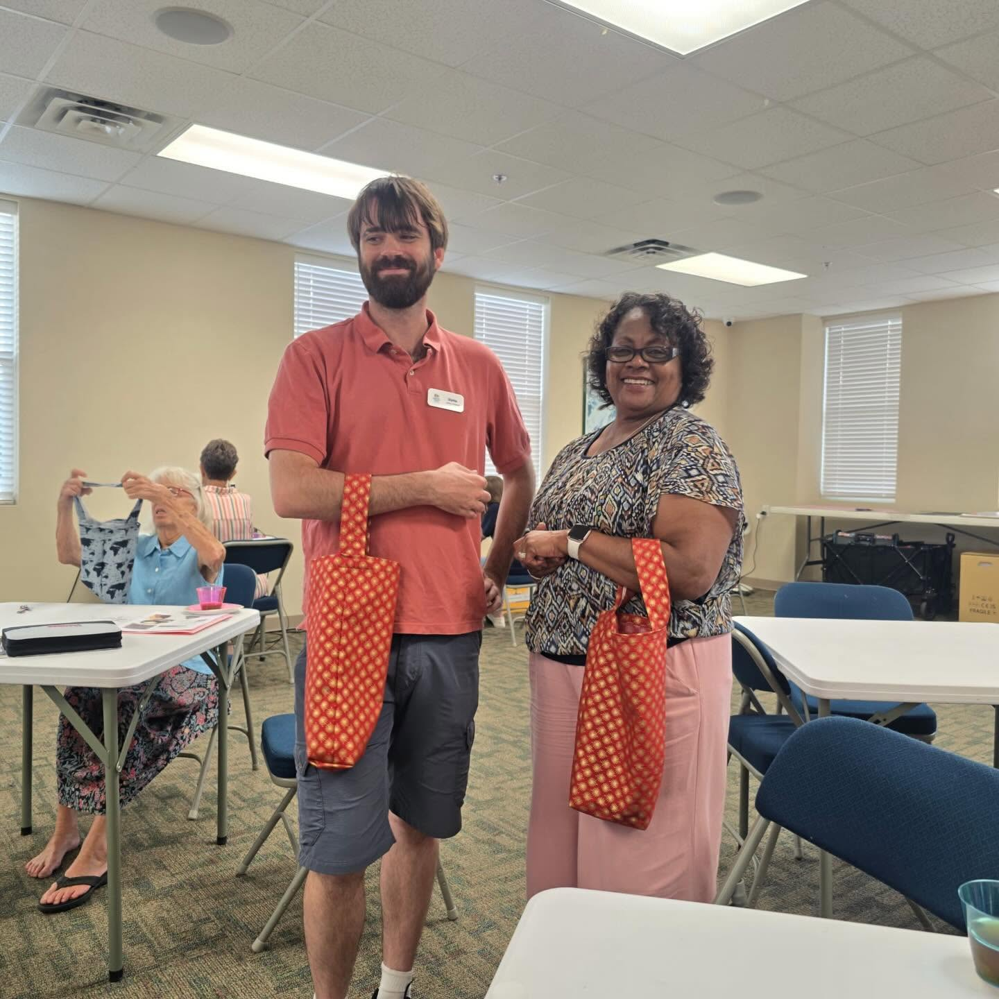 Man in orange shirt and blue shorts and woman in pink slacks and print top holding handmade tote bags in the library