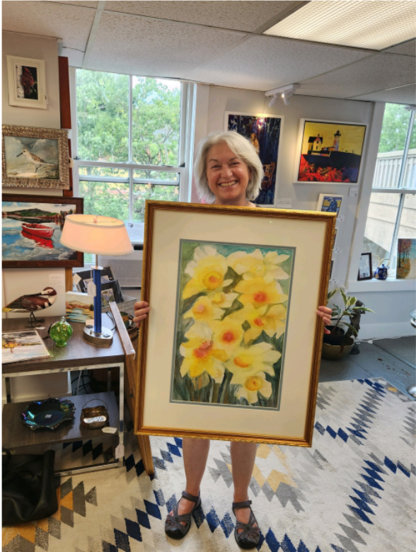 December artist Kathy Flygare holding up a watercolor painting of yellow flowers on a green background. December artist Kathy Flygare holding up a watercolor painting of yellow flowers on a green background.