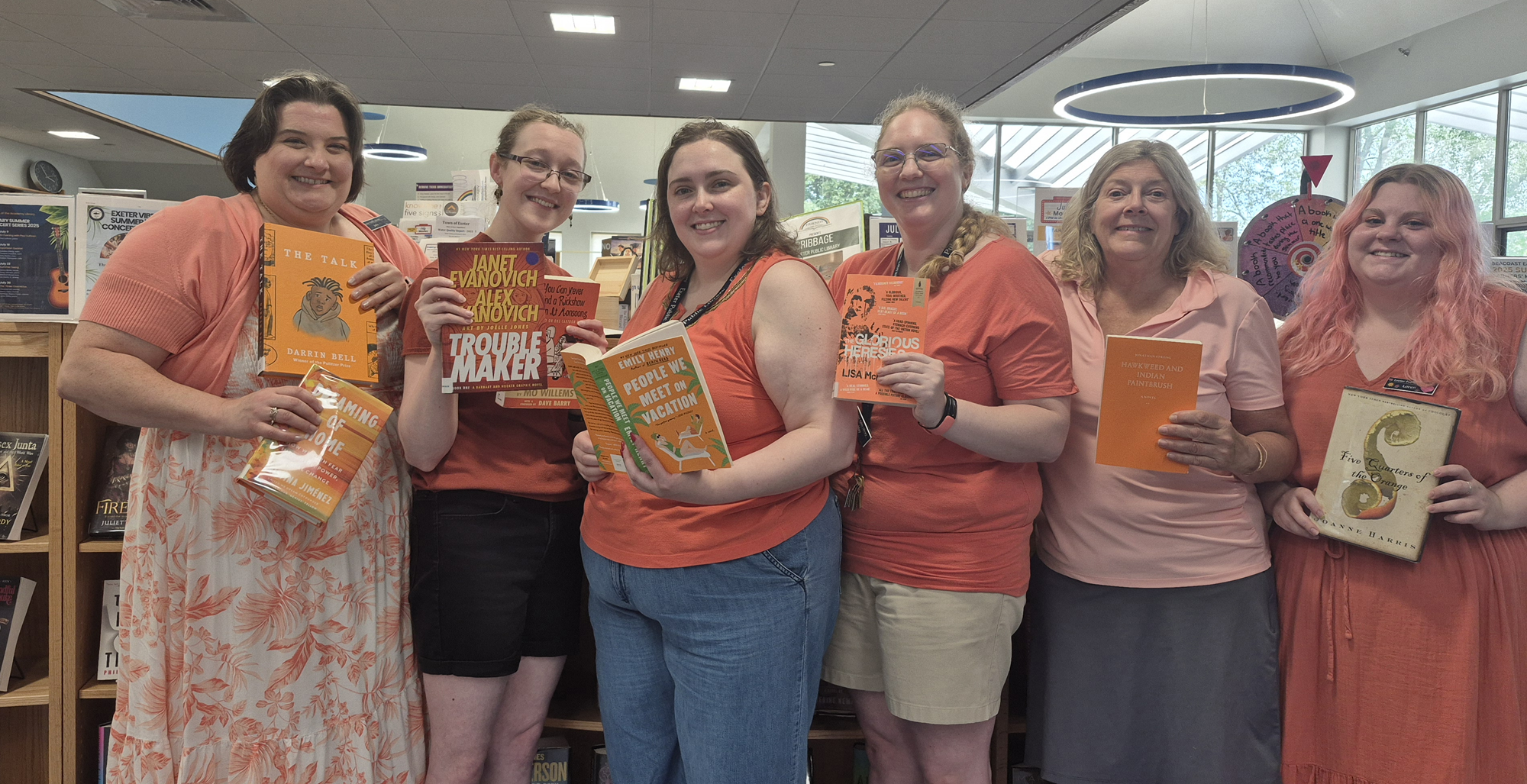 Staff members posing with books in front of the new book shelf on the main floor of the library. Staff members posing with books in front of the new book shelf on the main floor of the library.