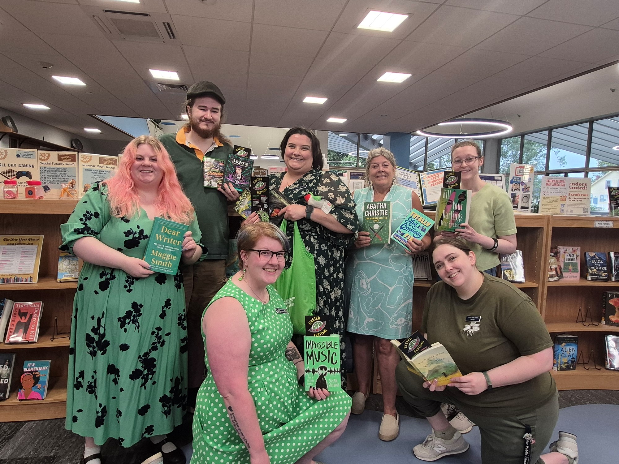 Staff members wearing green and posing with books in front of the new book shelf on the main floor of the library.