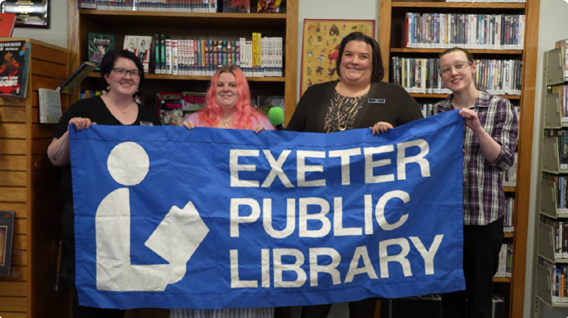 Happy Thanksgiving from Exeter Public Library! Four staff members hold "Exeter Public Library" banner in front of bookshelves.