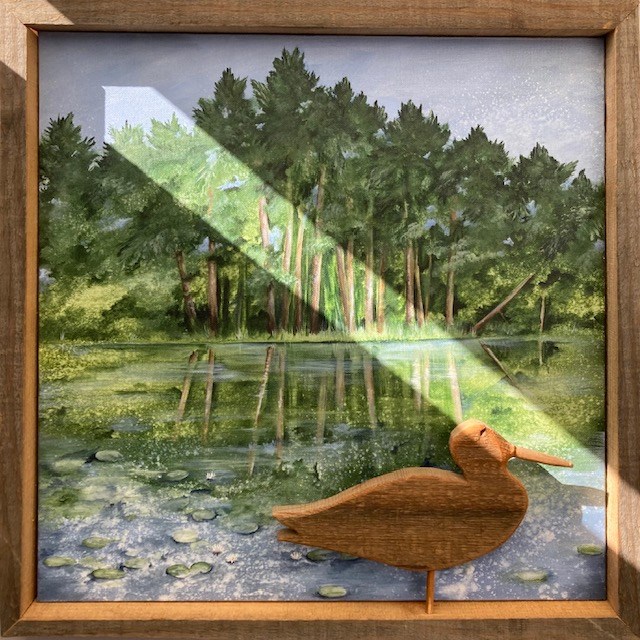 Painting of a lake with a forest behind it by April artist Nicole Ellis. The frame has a carved duck in the bottom right corner. A slice of sunlight runs diagonally across the painting from the top left corner to the bottom right. Painting of a lake with a forest behind it by April artist Nicole Ellis. The frame has a carved duck in the bottom right corner. A slice of sunlight runs diagonally across the painting from the top left corner to the bottom right.