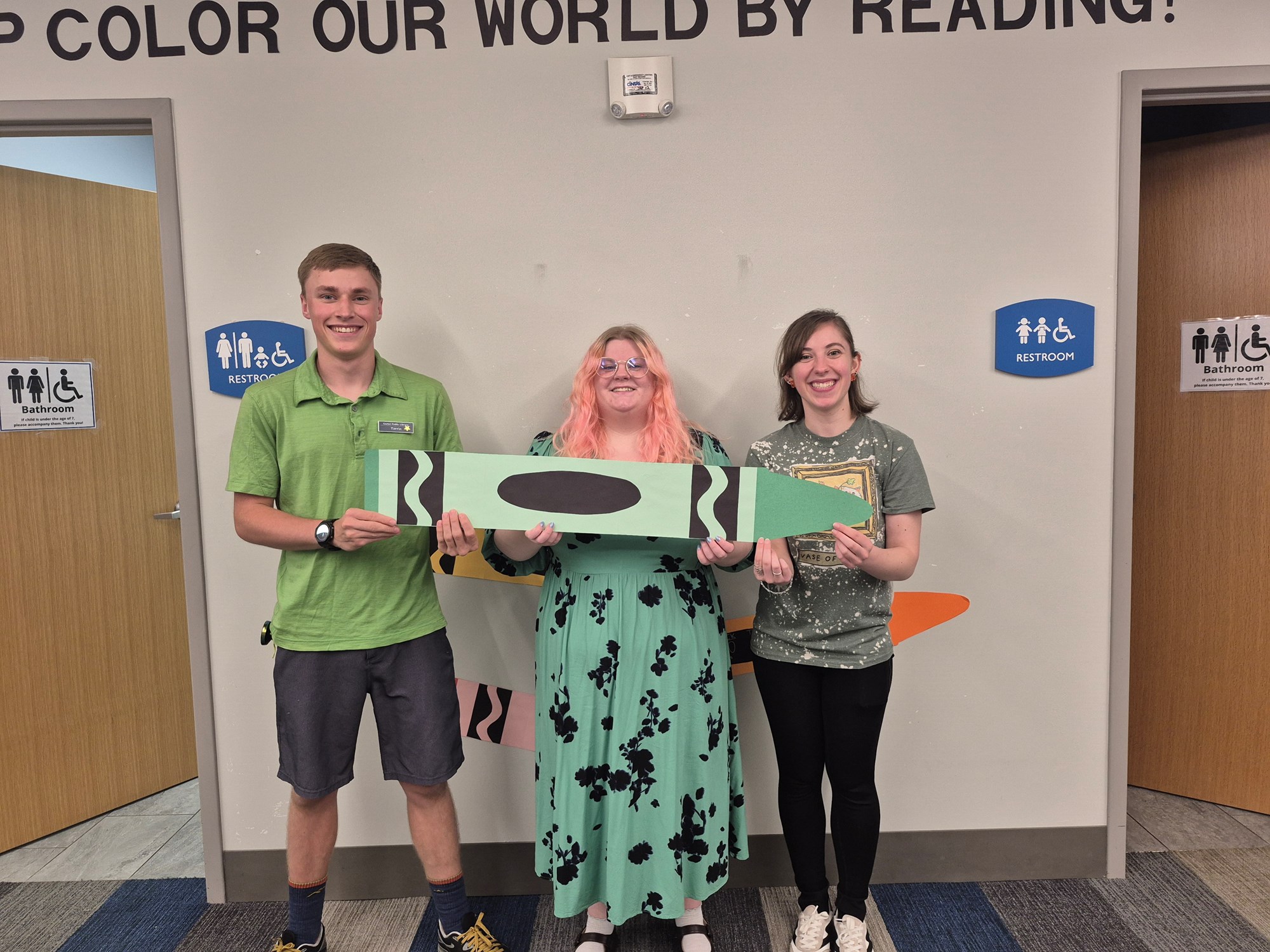 Staff members wearing green and posing with a giant green crayon in the Children's Room.