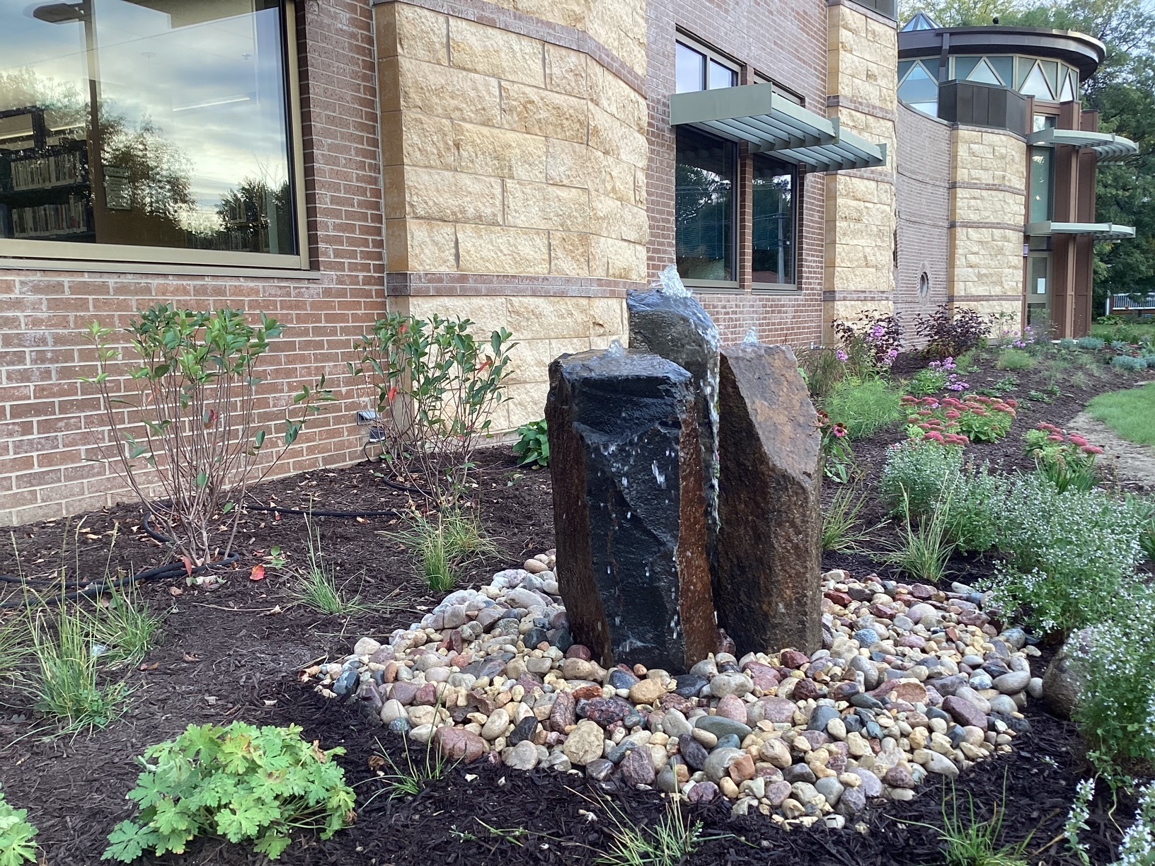 Three black basalt stone pillars surrounded by river rock in the middle of a plant bed next to the brick library building.