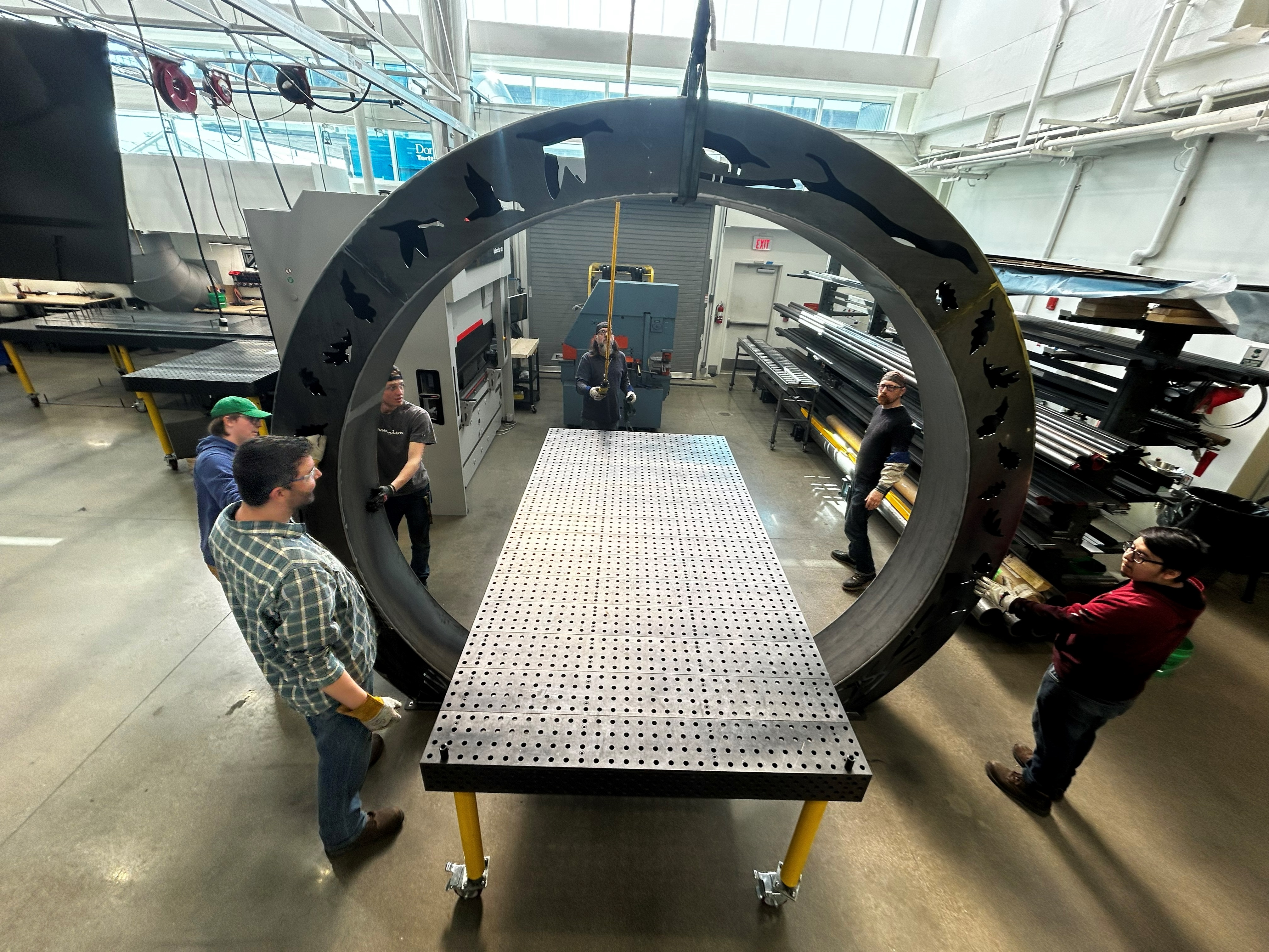 A large steel arch being hoisted by a crane. Cut-outs depicting native plants and wildlife are cut into the steel.