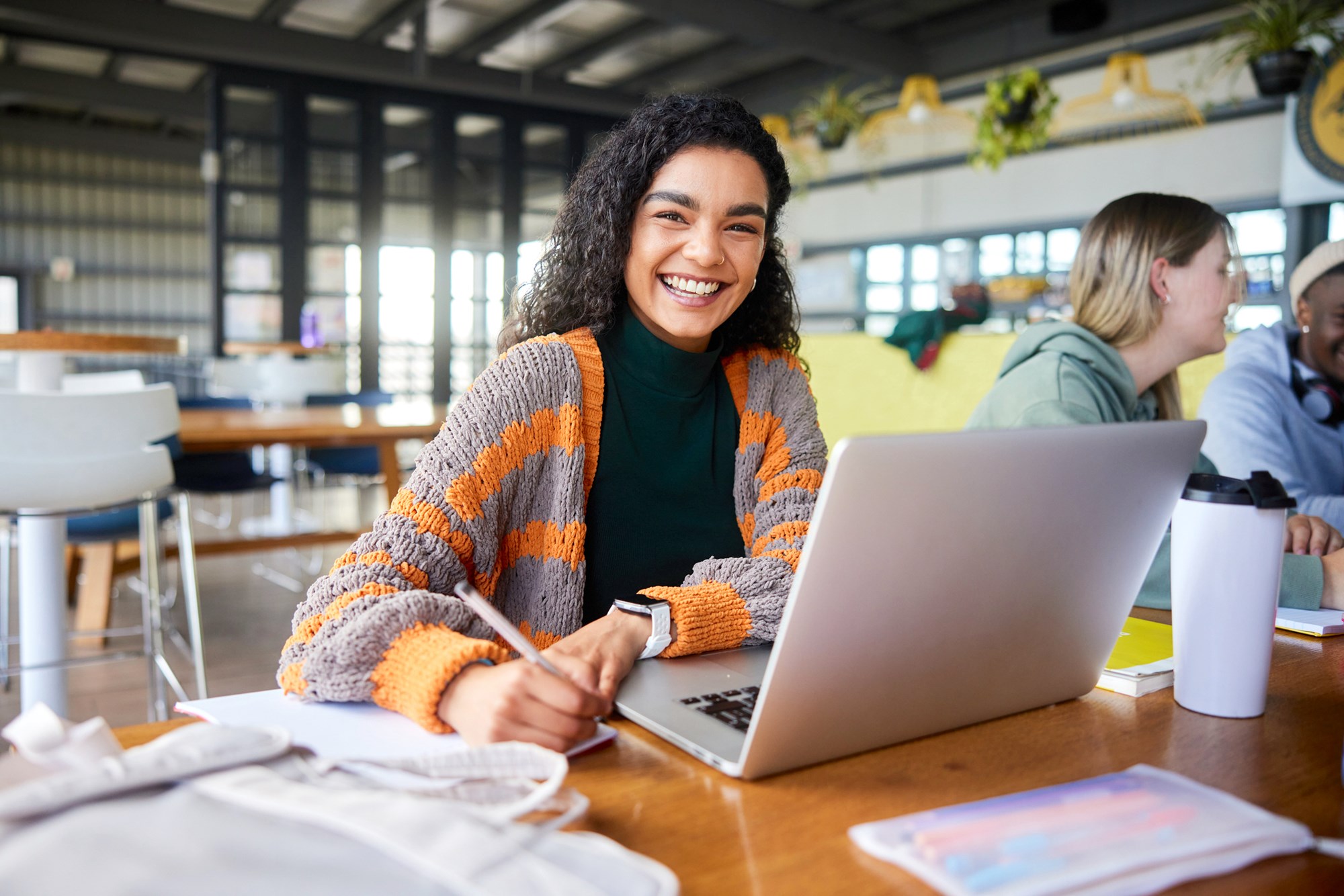 Young Female Student Laughs While Taking Notes and Working on her Laptop in a Campus Cafeteria