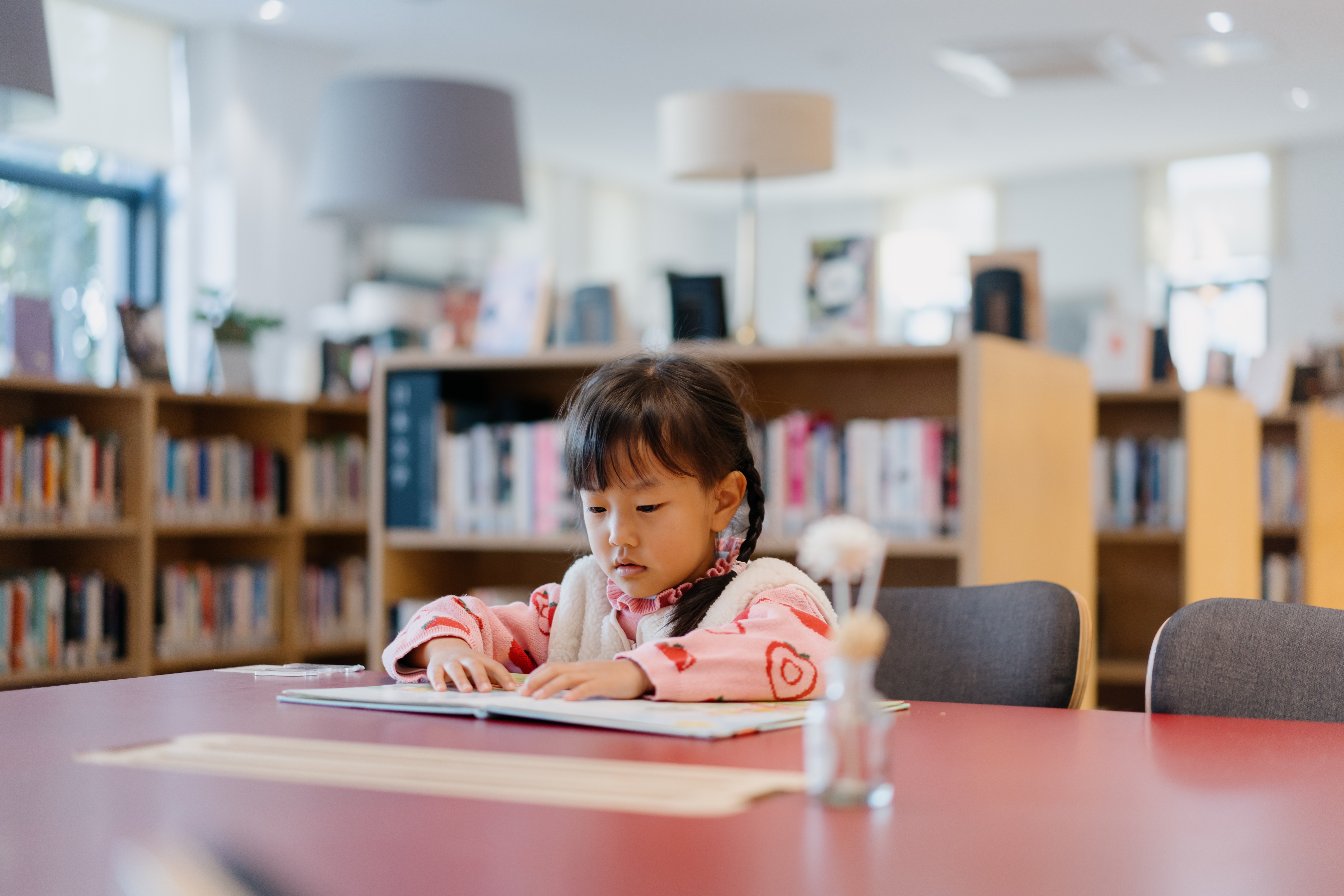 Little Girl Is Reading A Book On The Desk In The Library