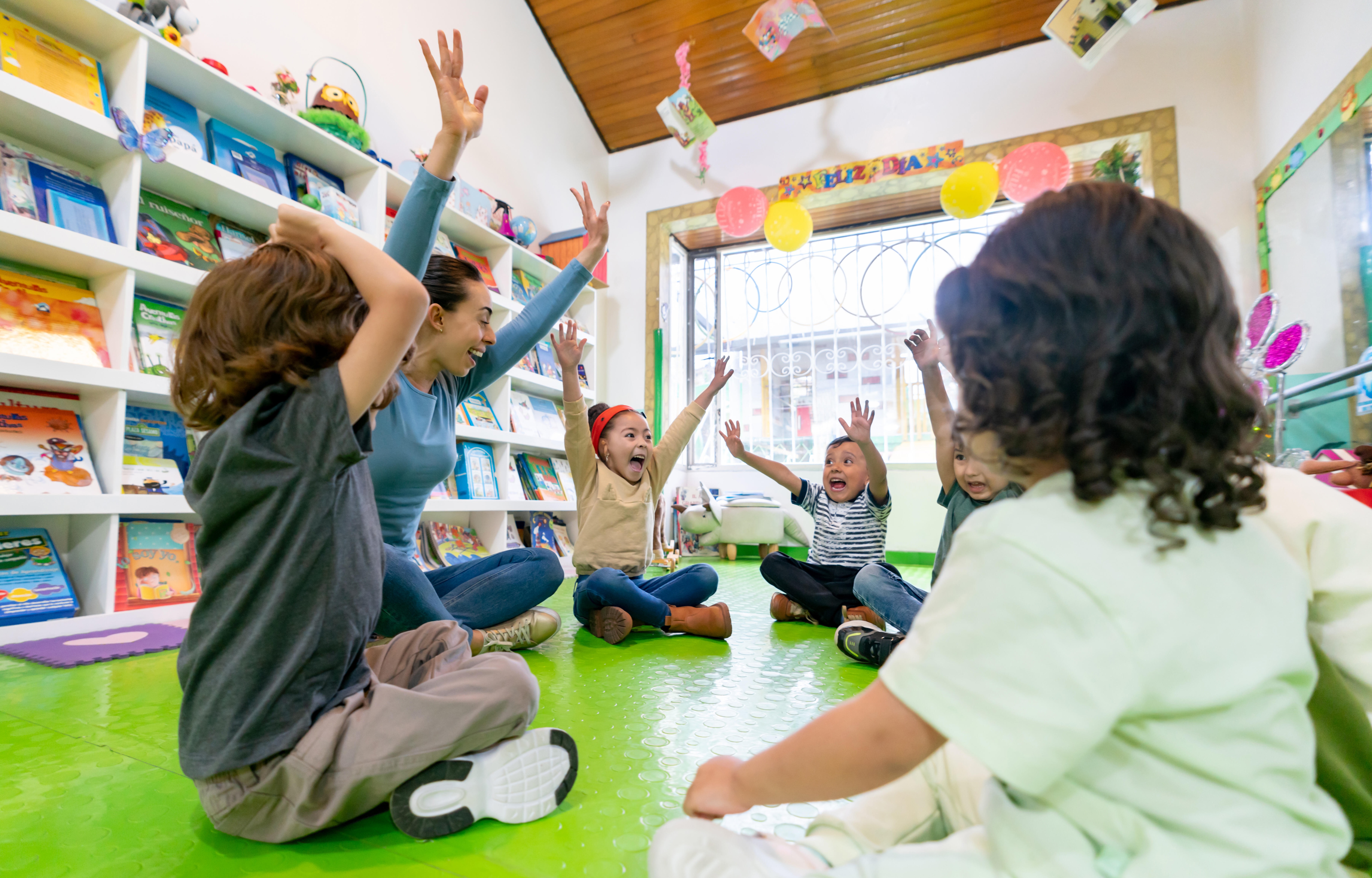 A group of children and a teacher sitting in a circle with their hands in the air. 