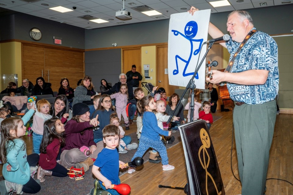 Russ performing for a group of children holding up a large drawing of a stick figure. 