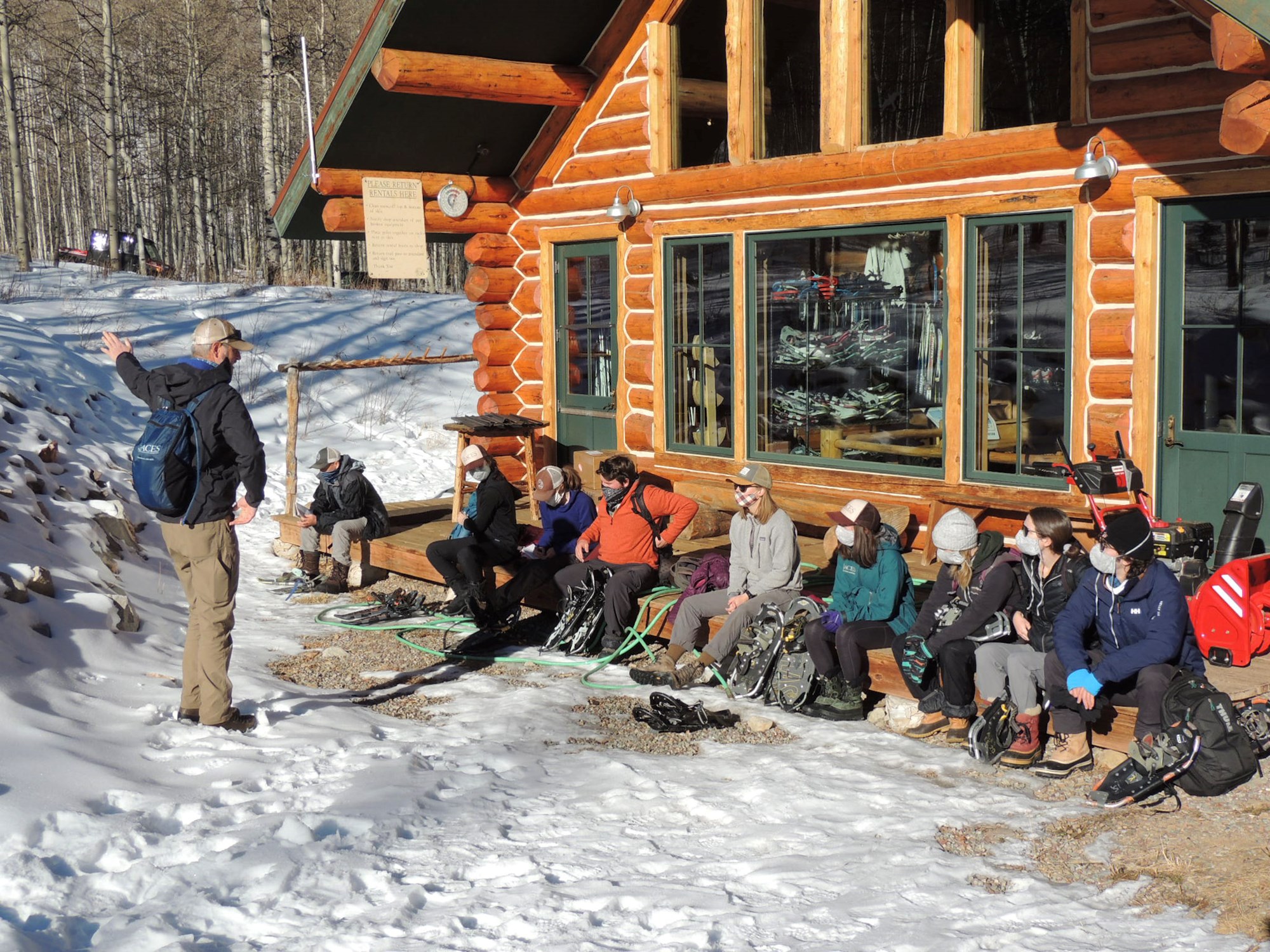 Ten people in winter gear listening to a presenter outside a cabin.