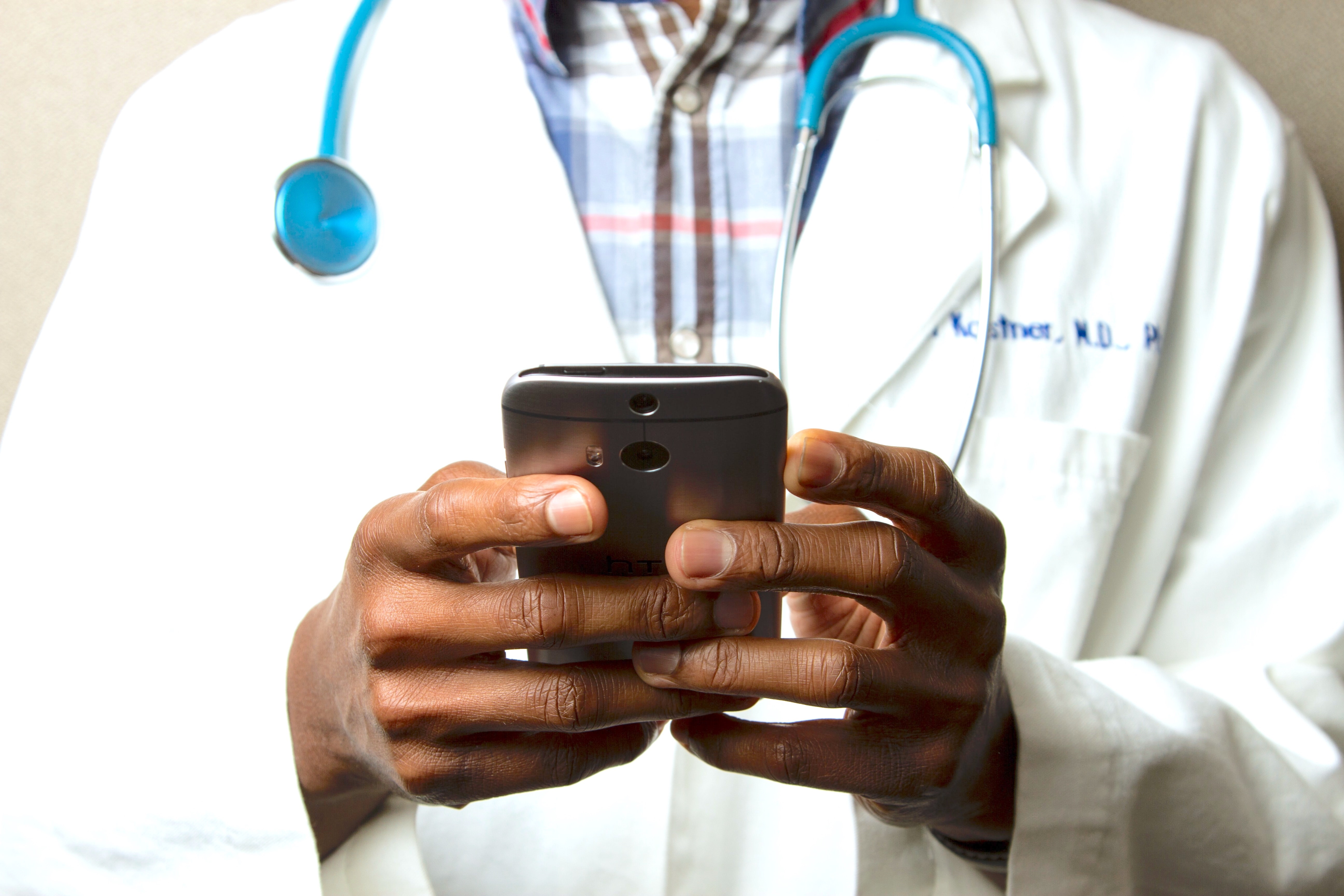 Image depicts the body of a doctor with a stethescope.  He is holding a phone in his hands.