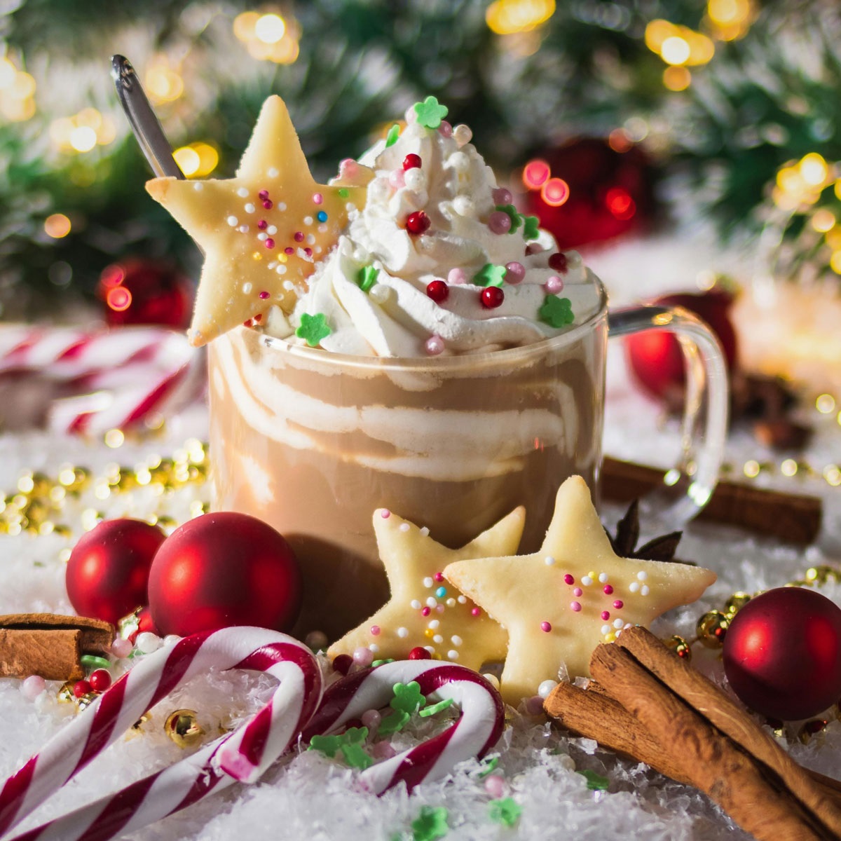 A mug of hot cocoa with whipped cream and sprinkles surrounds by candy canes and cookies