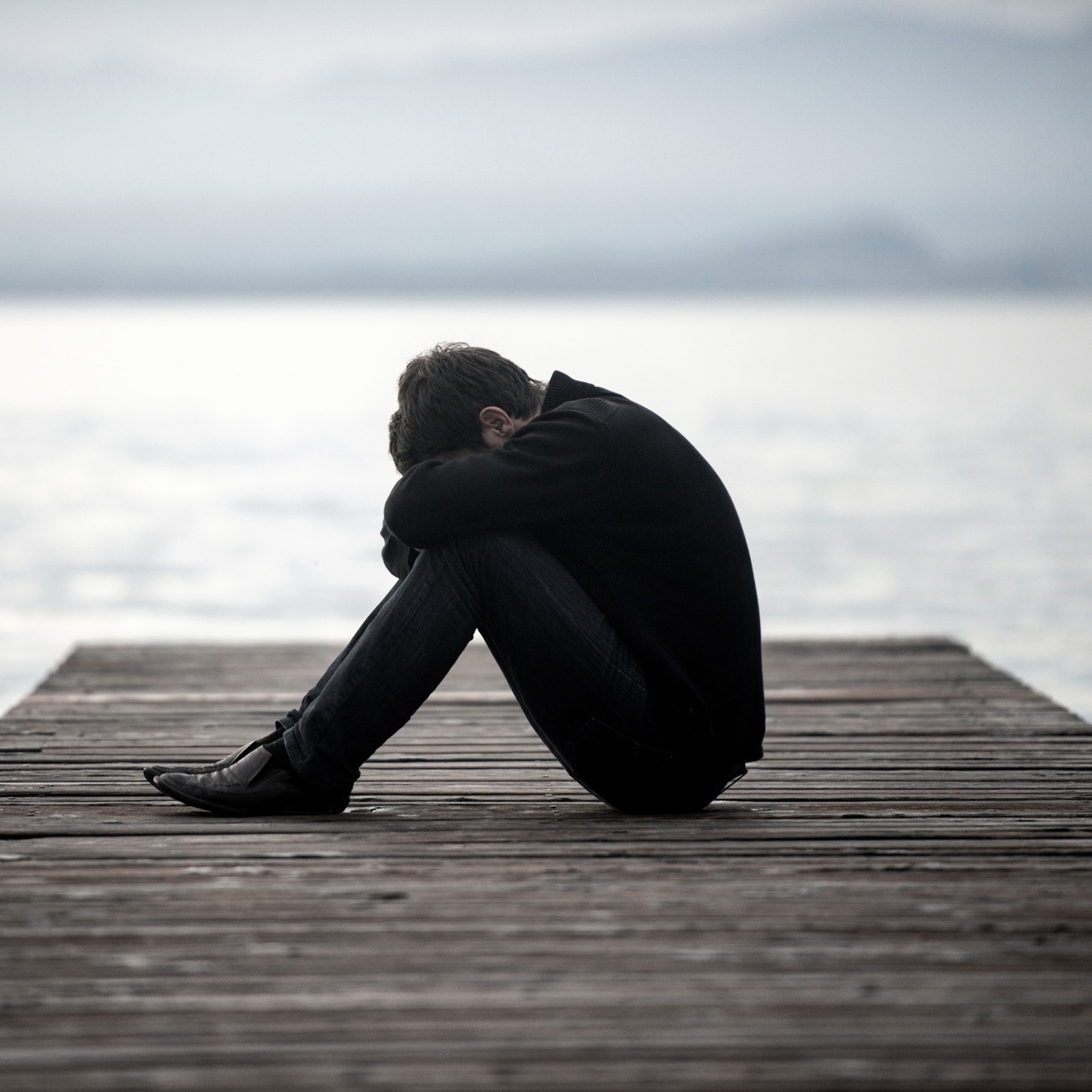 Overcast and gray waters, a person sits at the end of a dock with their head on their knees.