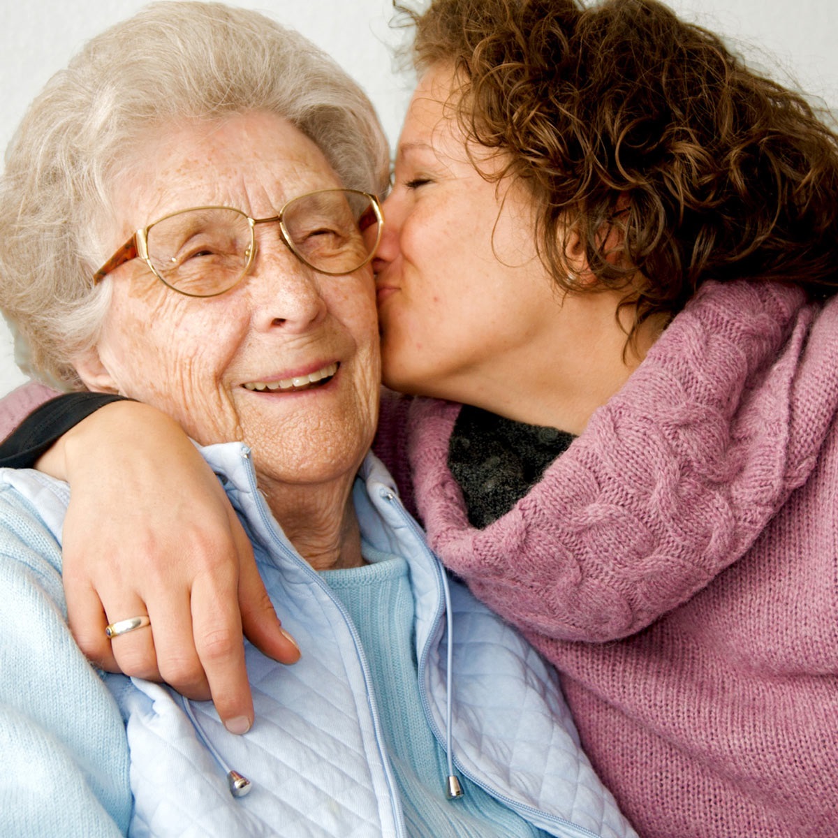Image shows a younger woman in a purple turtle neck kissing the cheek of an older woman who is wearing a blue fleece as well as glasses.