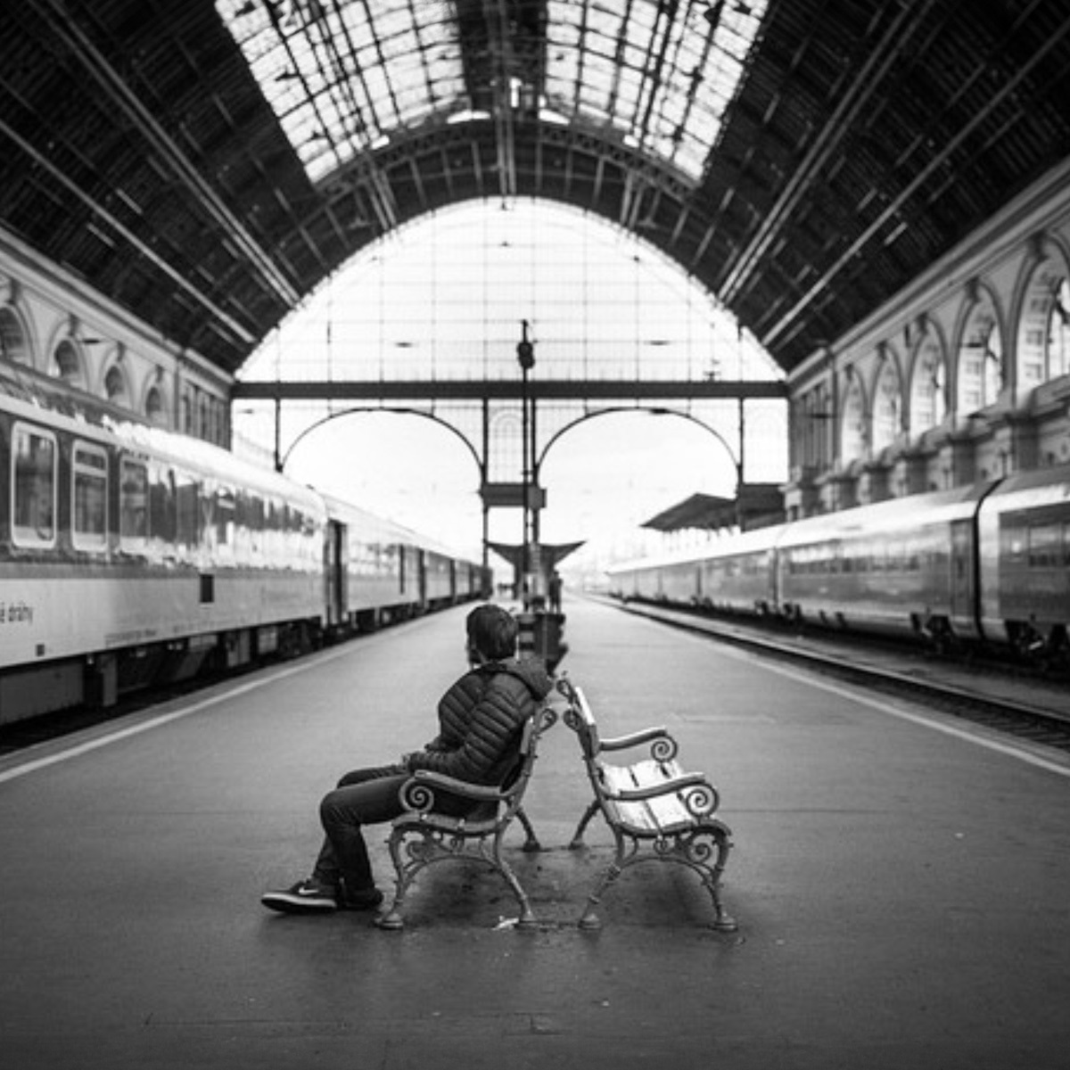 This is a black and white image of a train station platform.  There are trains on either side of the platform and a person sitting on a bench in the center area.