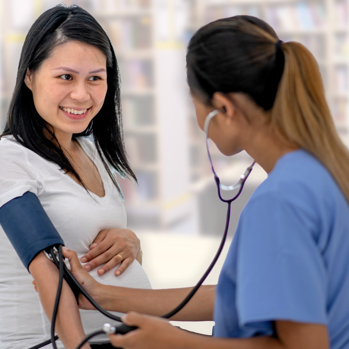Image is of a female in scrubs taking the blood pressure of a female in a white t-shirt.