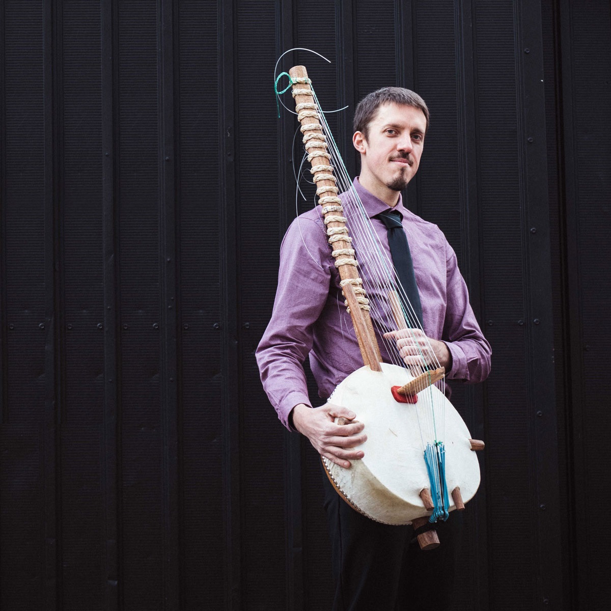 On a dark background Sean Gaskell holds a west african kora (a guitar-like instrument with a body built out of a gourd and a long neck).