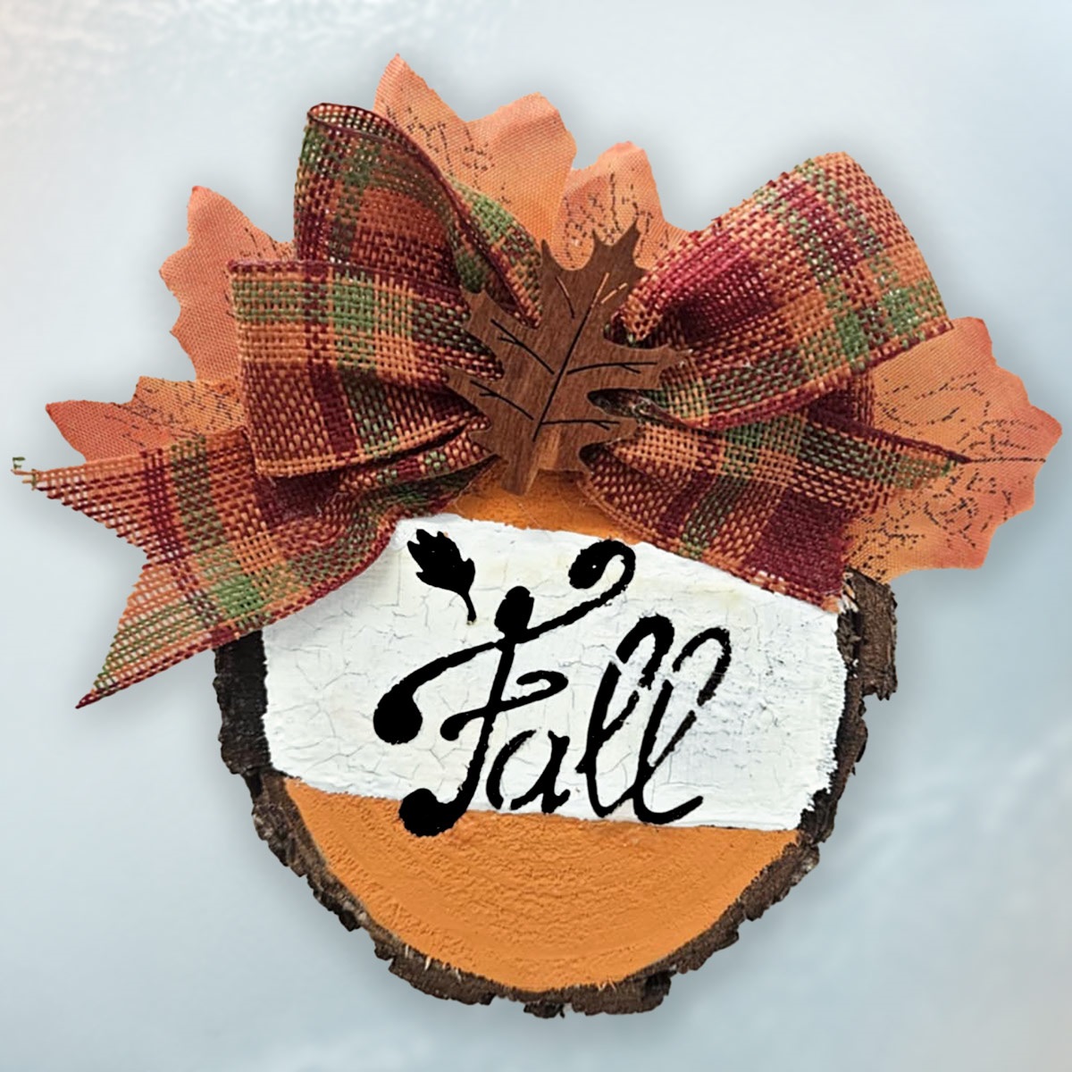 On a white background is small cross cut from a tree.  It is painted in fall colors, topped with fall colored bow and reads, "Fall" in fancy black script.