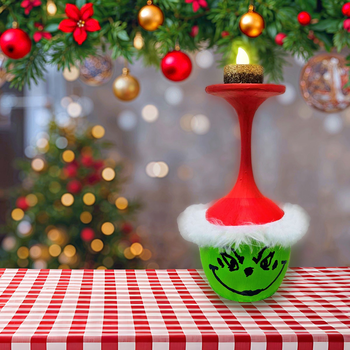 On a background of blurry christmas tree and christmas lights is a red and white checkered tablecloth.  On the table sets a wine glass votive painting to look like the grinch.