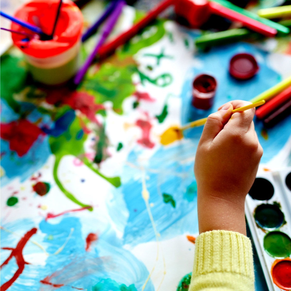 Lots of painting supplies on top of a painting a kids is working on.