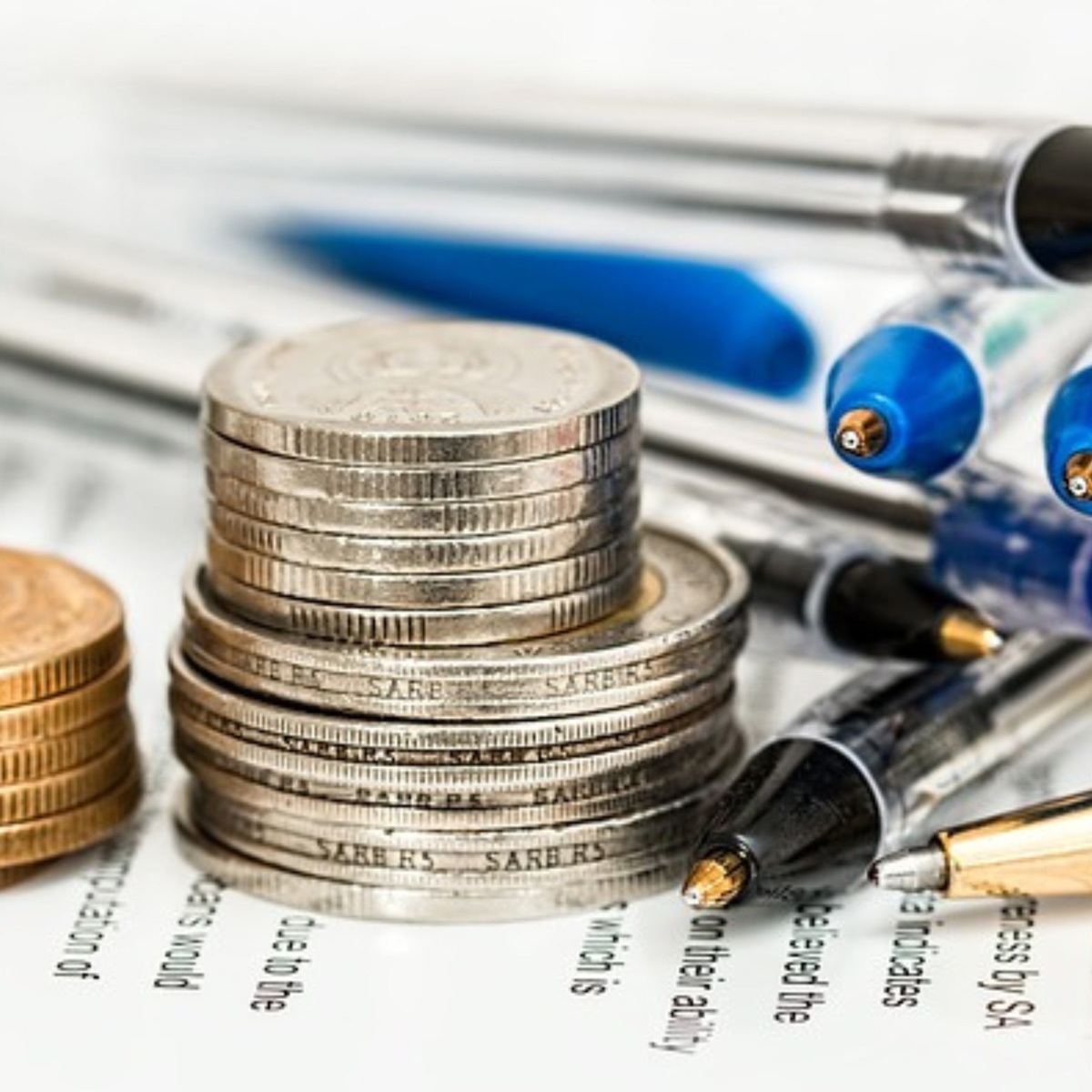 Image depicts small coin stacks and a bunch of pens on an unreadable text background.