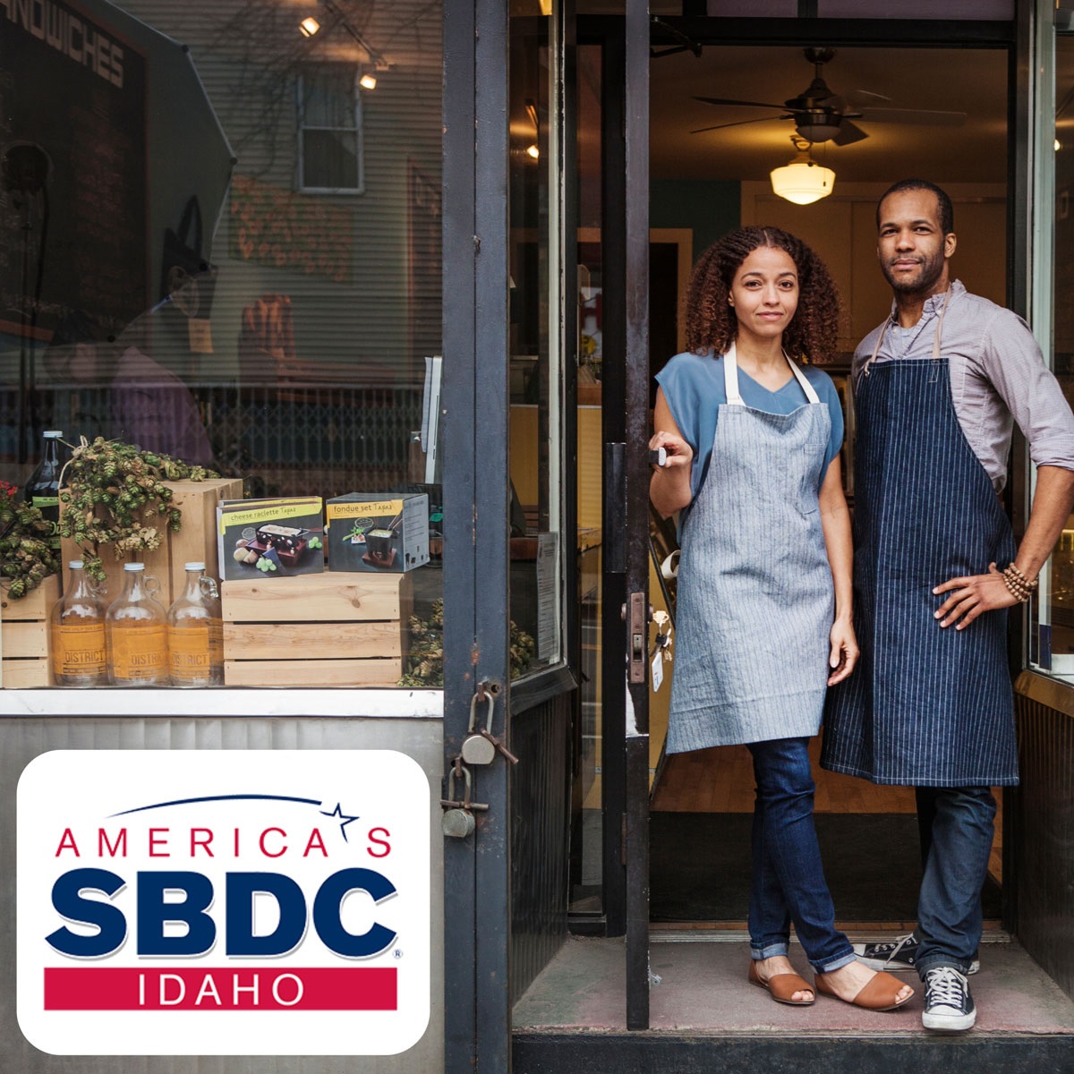 Image depicts a man and a woman in work aprons standing in the door of a shop.  On the bottom left corner their is a logo for America's SBDC Idaho.