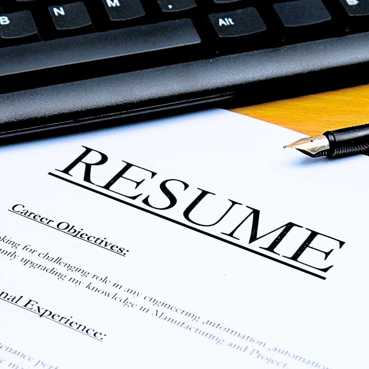 Images depicts a paper saying 'resume' in bold letters with the corner tucked under a computer keyboard on a wooden desk.