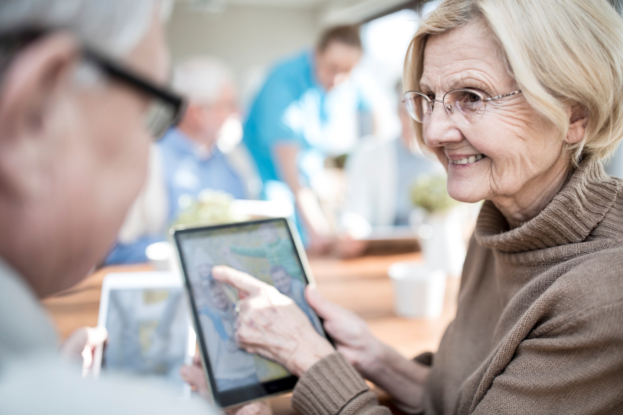Image depicts senior woman holding touch screen showing another senior something on the screen.