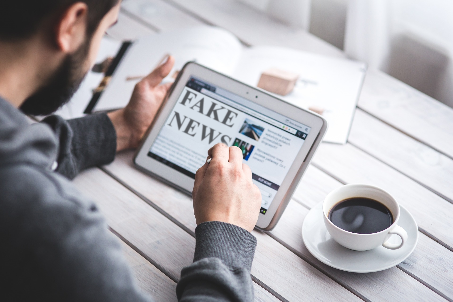 Image is a photograph of a man at a table holding an ipad with the words fake news on it.  There is a cup of coffee on the table.
