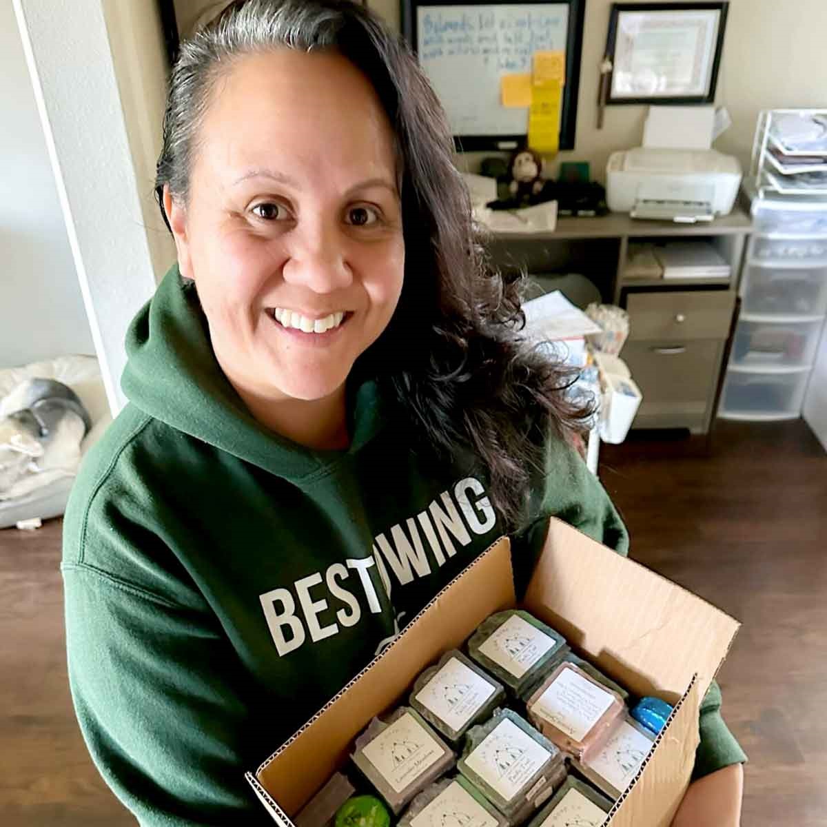 Image shows the smiling founder of Bestowing the Basics holding a box of what looks like soap bars.