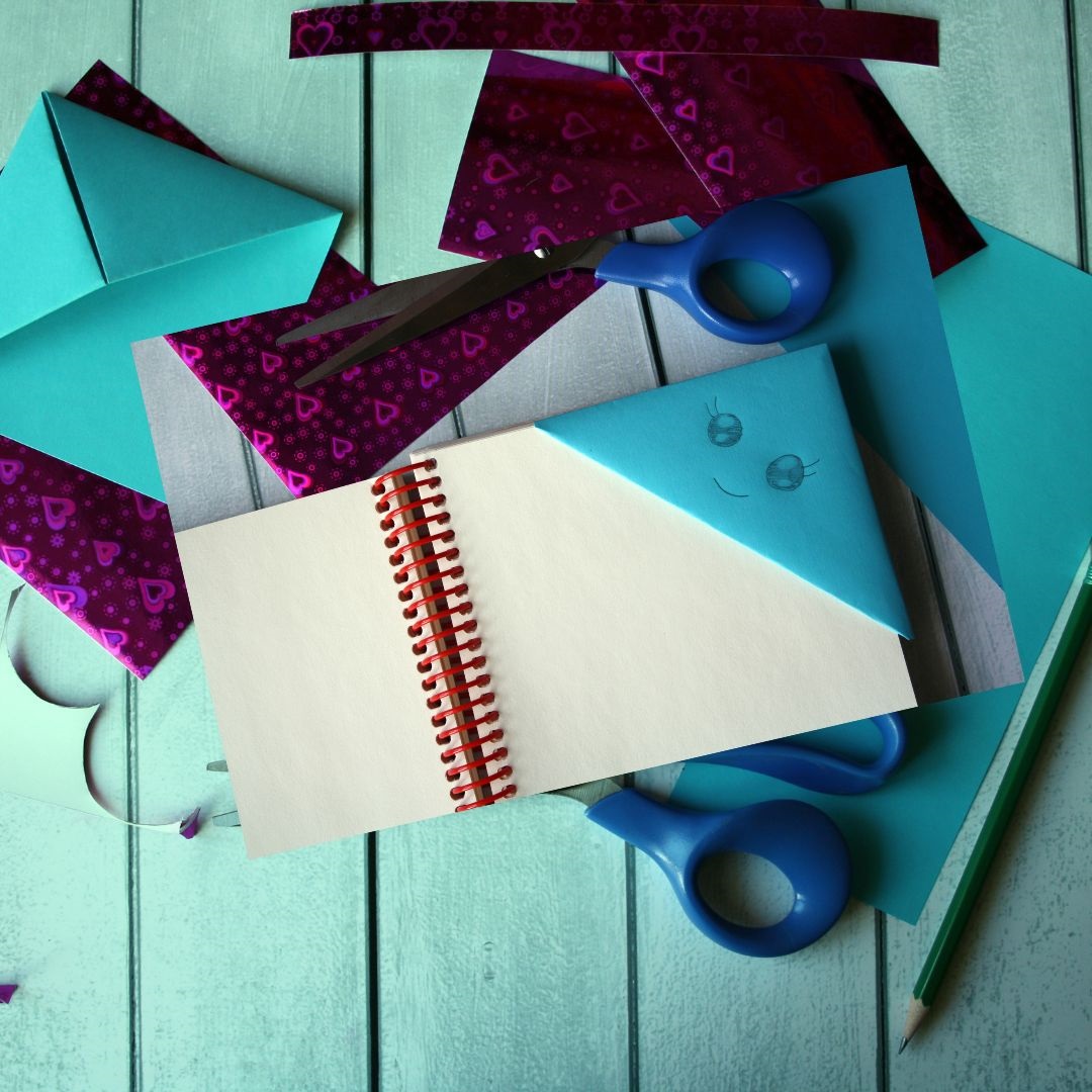 Image depicts a scattering of origami paper and scissors.  In the center there is a blank book with an origami bookmark folded around the top right corner.